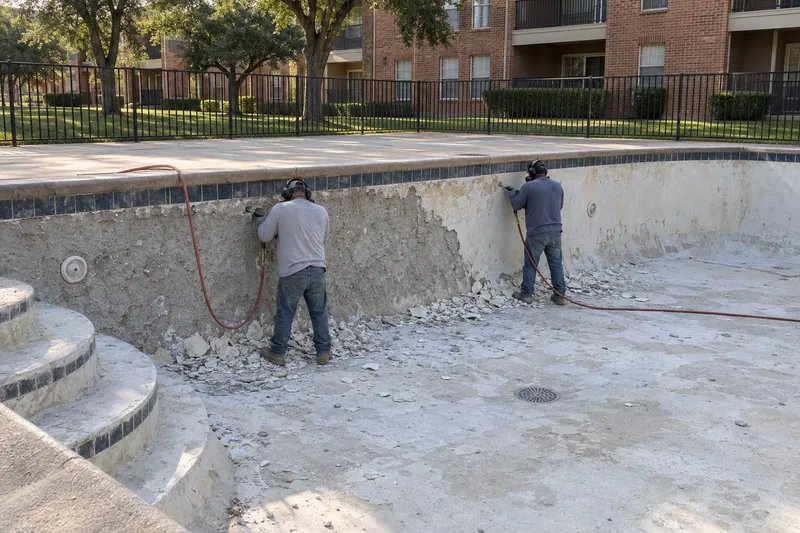 Old plaster being chipped off the wall of a large drained apartment pool